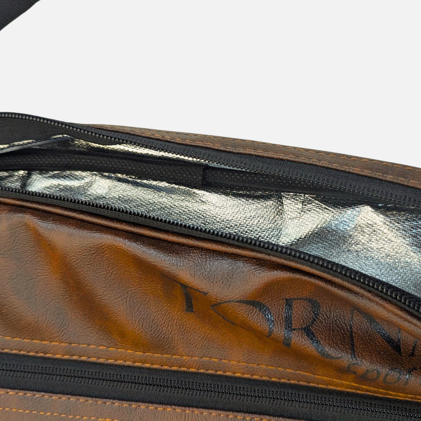 Close-up of a brown leather padel tennis racket cover and bag with black zippers and a reflective silver interior lining. Partial black logo text is visible on the leather surface.