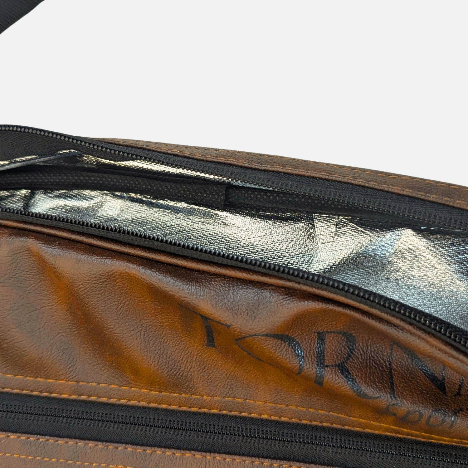 Close-up of a brown leather padel tennis racket cover and bag with black zippers and a reflective silver interior lining. Partial black logo text is visible on the leather surface.